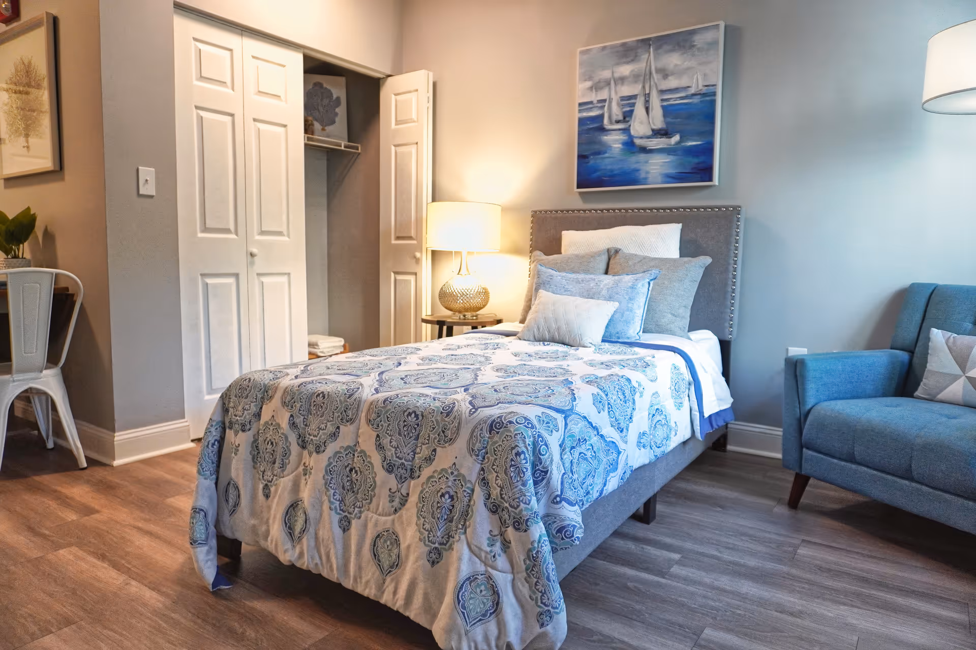 A cozy bedroom with a single bed covered in a blue and white patterned quilt and multiple pillows. There is a nightstand with a lamp beside the bed, a blue armchair with a decorative pillow, and a closet with white double doors. A painting of sailboats on the water hangs on the wall above the bed.