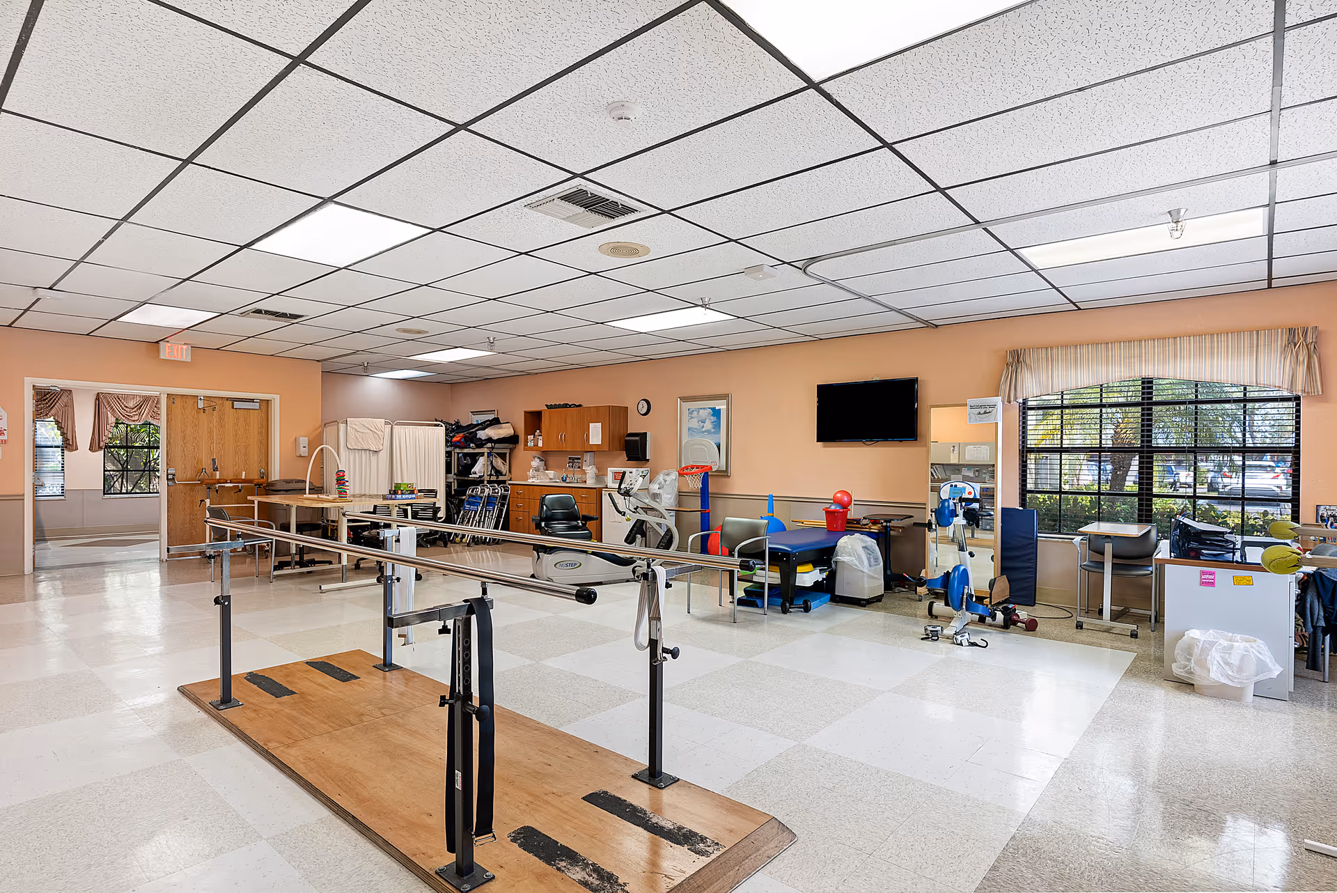 A spacious physical therapy room in a senior living facility with parallel bars for walking exercises, exercise bikes, therapy tables, and various rehabilitation equipment. The room has a tiled floor, peach-colored walls, a drop ceiling with fluorescent lights, and large windows letting in natural light.
