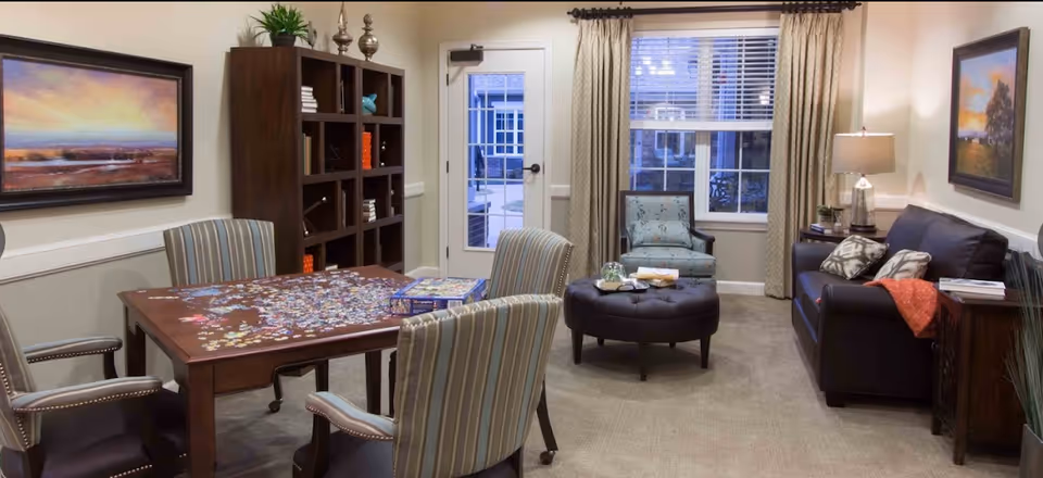 A cozy living room area with a wooden table covered in a partially completed jigsaw puzzle surrounded by four striped upholstered chairs. Behind the table is a tall dark wooden bookshelf with decorative items and books. To the right, there is a black leather sofa with patterned pillows and an orange throw, a side table with a lamp, and a framed landscape painting on the wall. Near the window, there is a cushioned armchair and a round ottoman with a glass cloche and books on top. The room has beige walls, carpeted floor, and a door with a window leading outside.