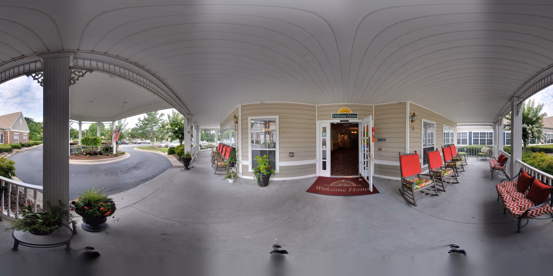 Covered entrance porch of Oaks at Evans facility with a 'Welcome Home' mat and sign above the door. The porch has several red cushioned rocking chairs and benches with red and white patterned pillows. There are potted plants along the walkway and a circular driveway with landscaping and trees visible outside.