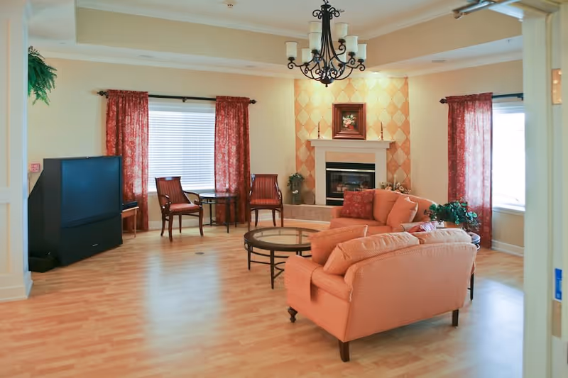 A cozy living room with light wood flooring, a peach-colored sofa set with cushions, a round glass coffee table, two wooden chairs with red cushions, a large TV on a stand, and a fireplace with a decorative painting above it. The room has two windows with red patterned curtains and a black chandelier hanging from the ceiling.