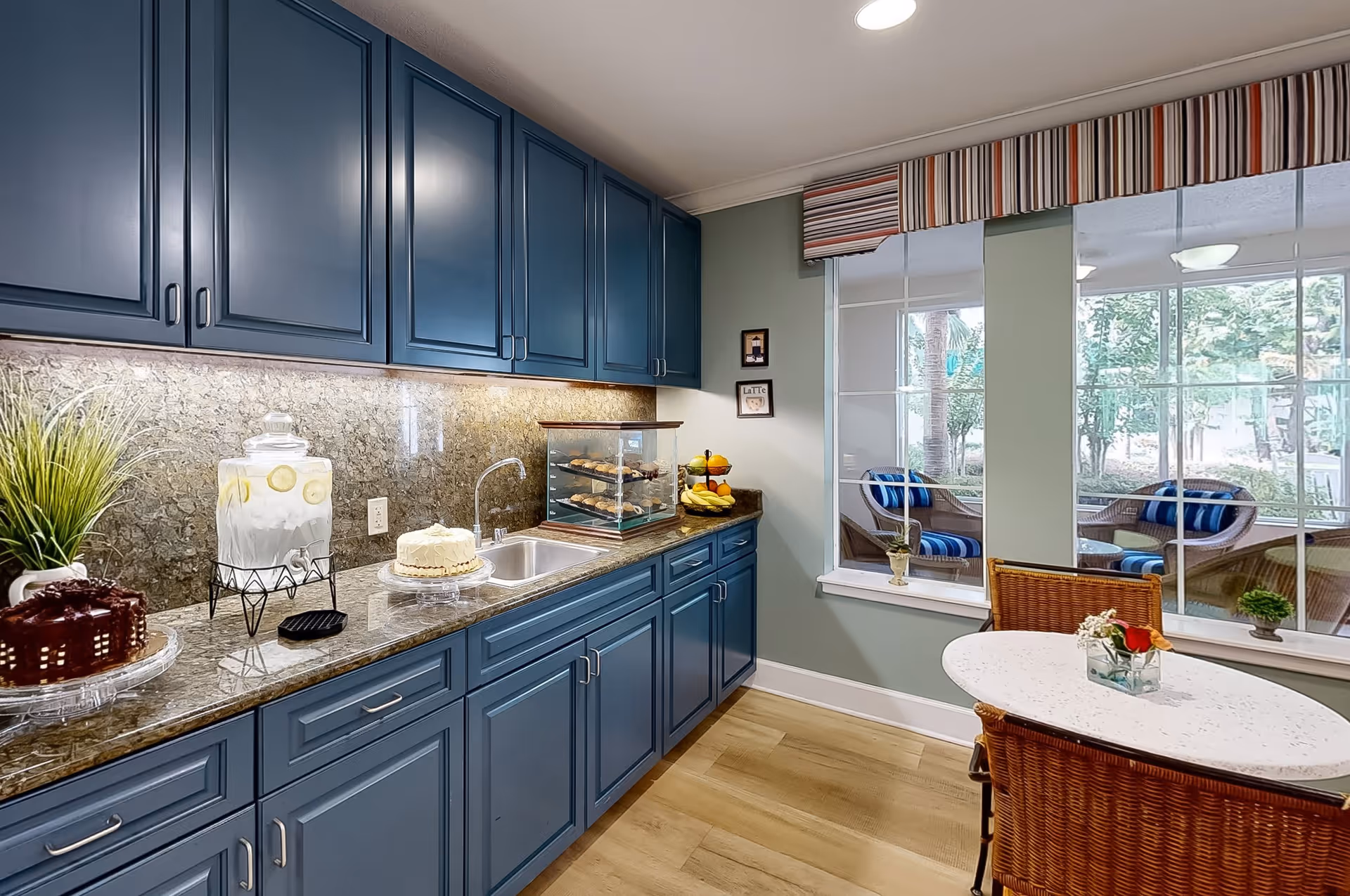 A bright kitchen area with blue cabinets and a granite countertop. On the counter, there is a glass container with lemon water, two cakes on stands, a small display case with pastries, and a fruit basket. A round table with two wicker chairs is placed near large windows that show an outdoor seating area with wicker chairs and blue cushions. The walls are painted light green and there is a striped valance above the windows.