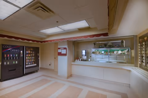 Interior view of a senior living facility area featuring vending machines for cold drinks and snacks on the left, a bulletin board on the wall, and a serving counter with a glass display case containing dishes and cups on the right.