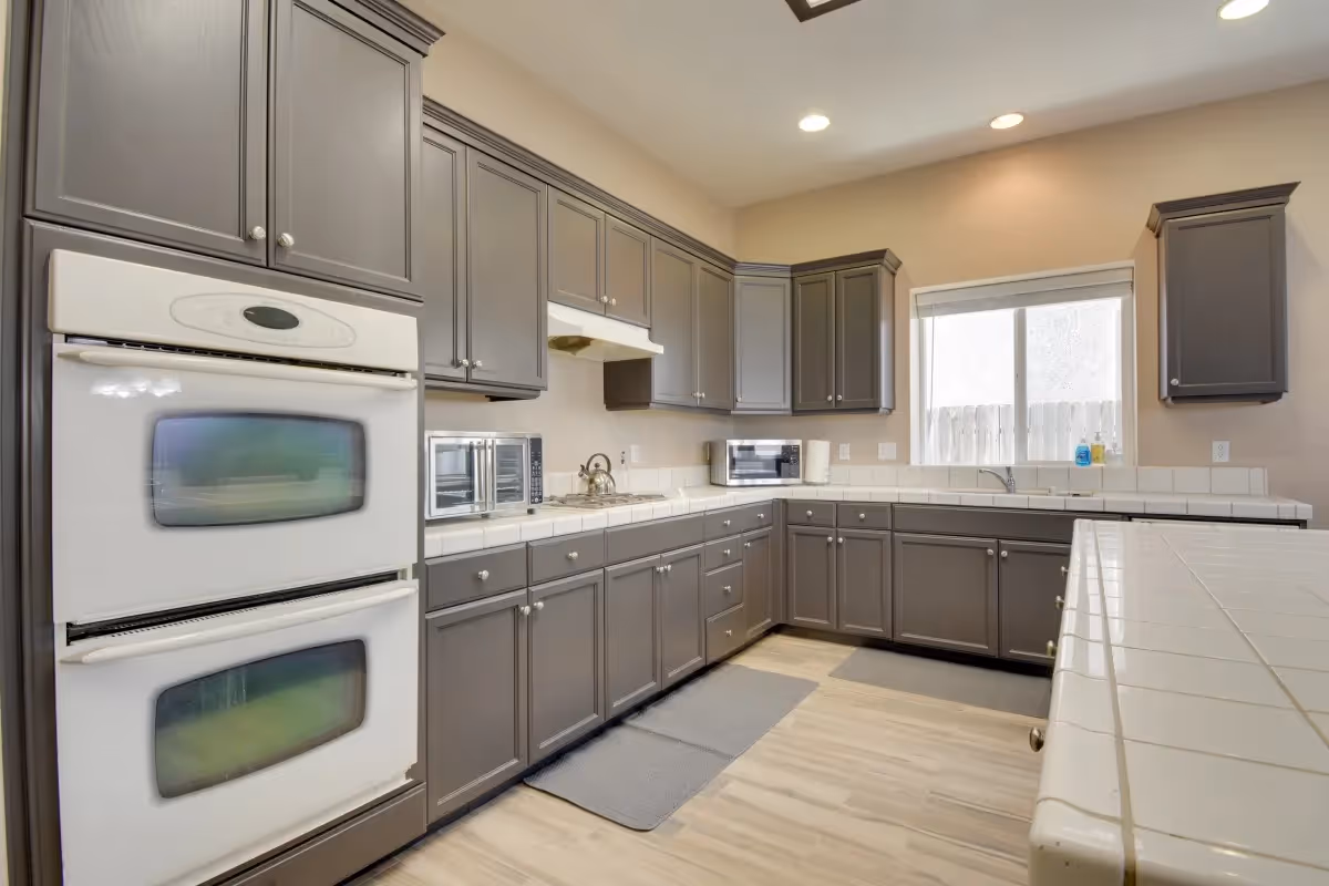 A modern kitchen with gray cabinets and white tiled countertops. The kitchen features a double oven, a gas stove with a kettle, two microwaves, a window above the sink, and light wood flooring with two gray floor mats.