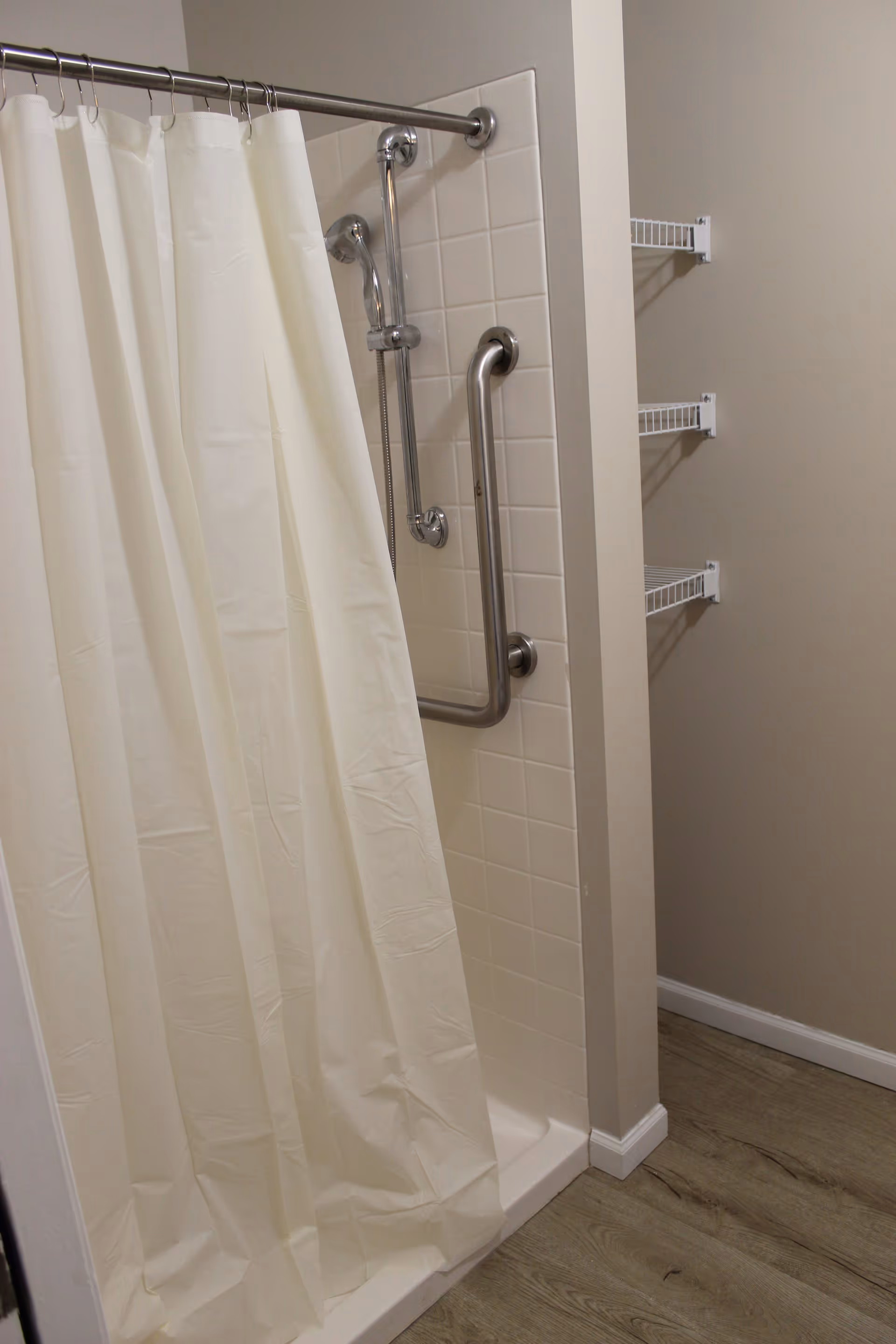A shower area with a white shower curtain, a metal grab bar, and a handheld showerhead mounted on a tiled wall. To the right, there are three white wire shelves mounted on a beige wall. The floor has wood-like vinyl flooring.