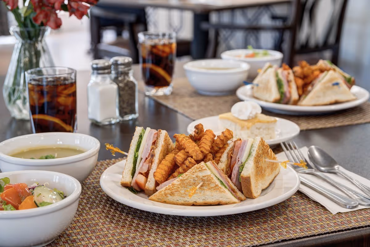 A dining table set with plates of club sandwiches with lettuce, tomato, and turkey, accompanied by crinkle-cut fries. There are bowls of soup and salad, glasses of iced tea with lemon slices, salt and pepper shakers, and a slice of cake with whipped cream on a small plate. The table has woven placemats and silverware wrapped in napkins.