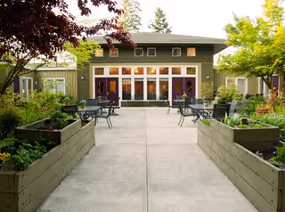 Courtyard with raised planter boxes and patio tables and chairs leading to a single-story building with large glass doors and windows.
