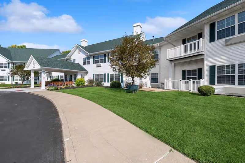 Exterior view of Park Square Manor, a two-story white building with green shutters and a green roof. There is a covered entrance with chairs underneath, a well-maintained lawn, a tree, and a curved sidewalk leading to the entrance. The sky is clear with a few clouds.