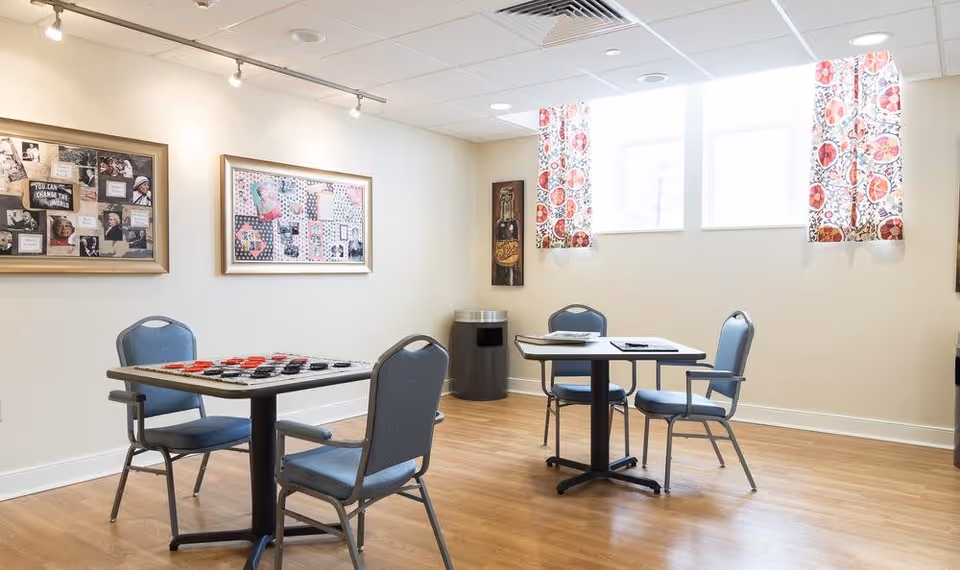 A bright room with two tables and four chairs arranged around them. One table has a checkers game set up, and the other has some papers on it. The room has light-colored walls, wooden flooring, two windows with colorful patterned curtains, framed pictures on the walls, and a trash can in the corner.