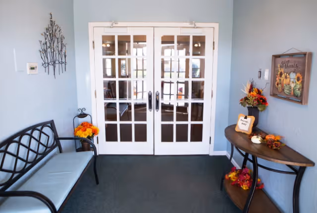 Small entryway with double glass doors leading to another room. On the left side, there is a black metal bench with a light blue cushion and a small pot of orange flowers beside it. On the right side, there is a wooden console table decorated with fall-themed flowers and a small sign. A framed artwork with sunflowers and the words 'give thanks' hangs on the wall above the table.