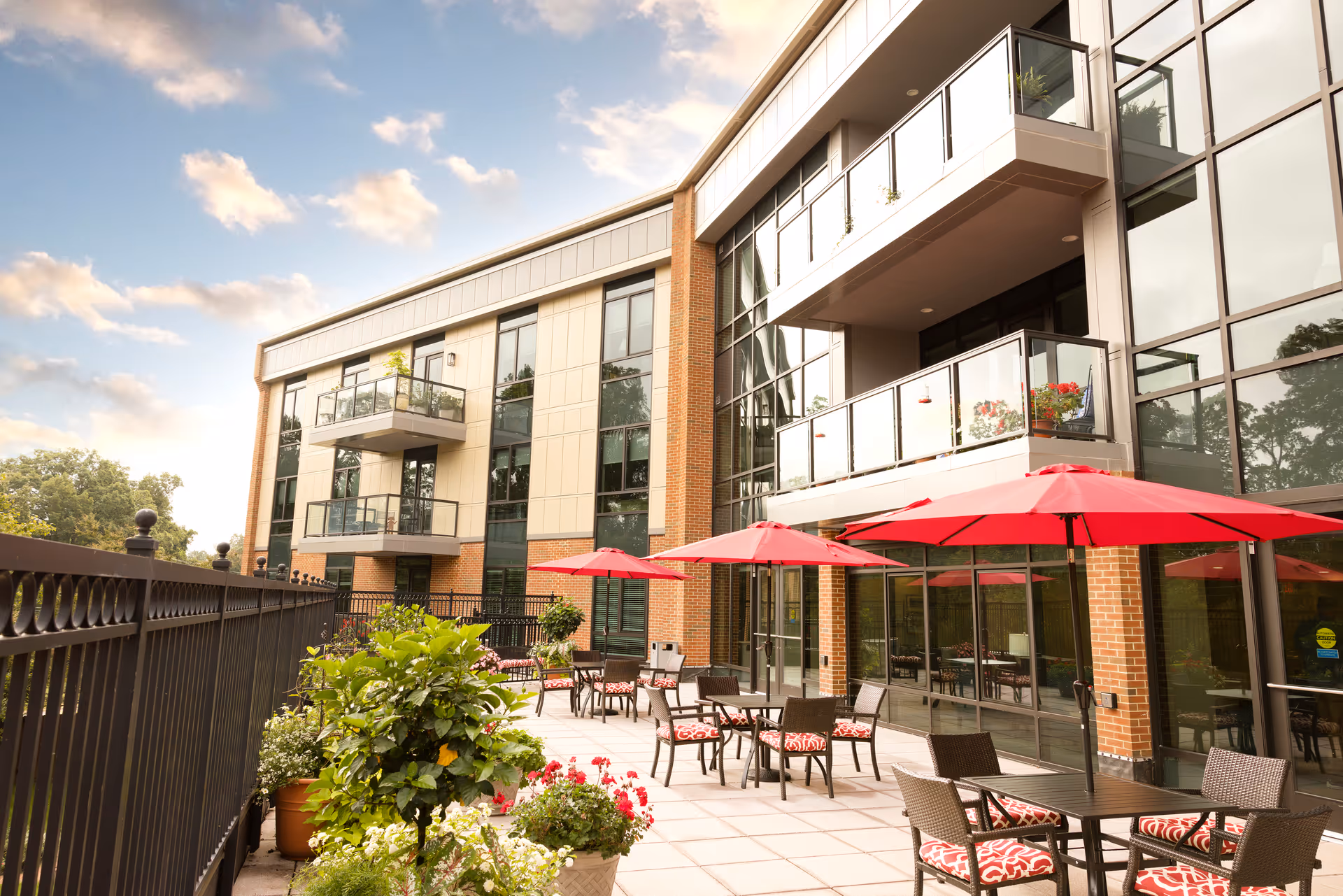 Outdoor patio area at Westminster Canterbury featuring tables with red umbrellas and cushioned chairs, surrounded by potted plants and flowers, adjacent to a modern building with balconies and large windows under a partly cloudy sky.