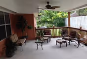 Covered outdoor patio area with cushioned seating including chairs and a loveseat, a glass-top coffee table, potted plants, ceiling fan, and a wooden railing overlooking a green backyard with trees and a wooden fence.