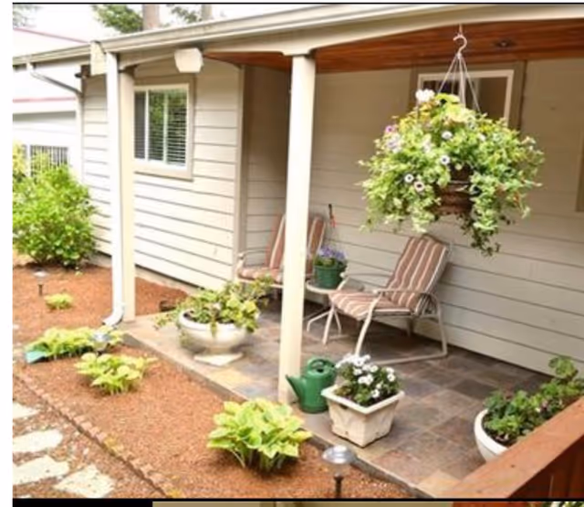 A covered patio area with two striped cushioned chairs and a small round table between them. The patio is surrounded by potted plants, including a hanging basket with flowers, and garden beds with green plants. The exterior wall of the building is light-colored with a window and a door visible.