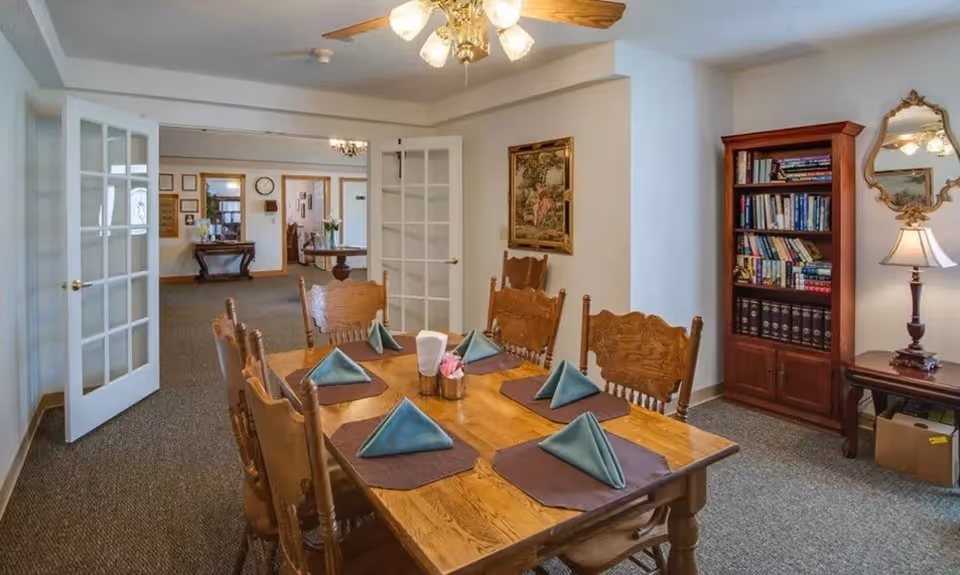 A dining room with a wooden table set with folded napkins and placemats, surrounded by carved chairs, a bookcase, and French doors opening to a hallway.