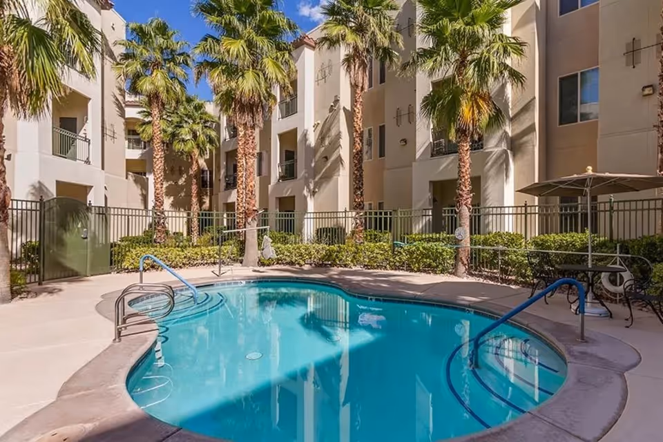 Outdoor swimming pool surrounded by palm trees and a multi-story residential building with balconies. There is a metal fence around the pool area and a table with an umbrella on the right side.