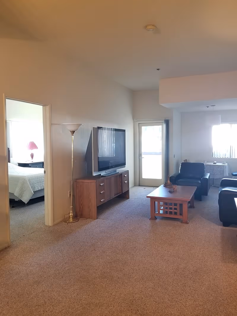 Interior view of a living room in a senior living facility with beige carpet, a wooden TV stand with a flat-screen TV, a floor lamp, a wooden coffee table, and black leather chairs. A doorway on the left leads to a bedroom with a bed and a bedside table with a pink lamp. A glass door and window with vertical blinds are visible in the background.
