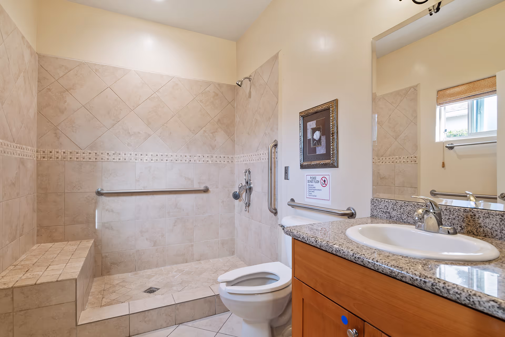 A spacious bathroom with beige tiled walls and floor, featuring a walk-in shower with a built-in bench and grab bars, a toilet, and a wooden vanity with a granite countertop and a sink. A large mirror is mounted above the vanity, and a small window with a blind is visible on the right side.