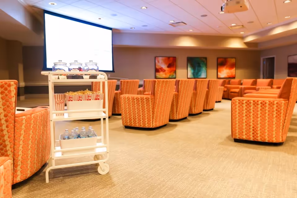 A cozy meeting or presentation room with multiple orange patterned armchairs arranged in rows facing a large projection screen. A white rolling cart in the foreground holds popcorn boxes, jars of candy, and bottled water. The room has warm lighting and colorful abstract paintings on the far wall.