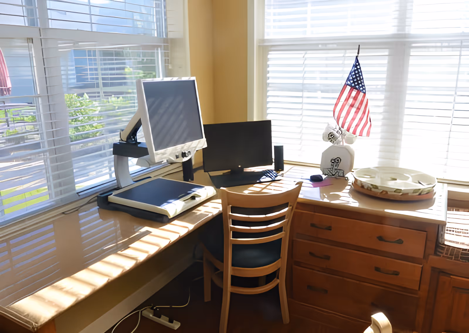 A sunlit corner office space with two computer monitors on a wooden desk next to large windows with white blinds. An American flag and some small decorative items are on the desk, along with a wooden chair in front of the desk.