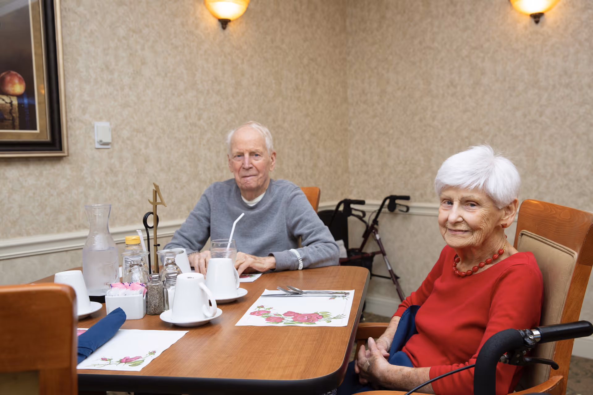 An elderly man and woman sitting at a wooden dining table in a room with beige wallpaper and wall sconces. The table is set with placemats, cups, a water pitcher, and condiments. A walker is visible behind the woman, who is wearing a red top and necklace, while the man is wearing a gray sweater.