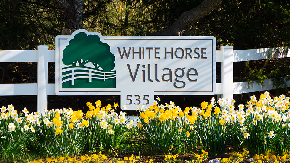 A white entrance sign reading "WHITE HORSE Village 535" mounted on a white fence with yellow and white daffodils in front.