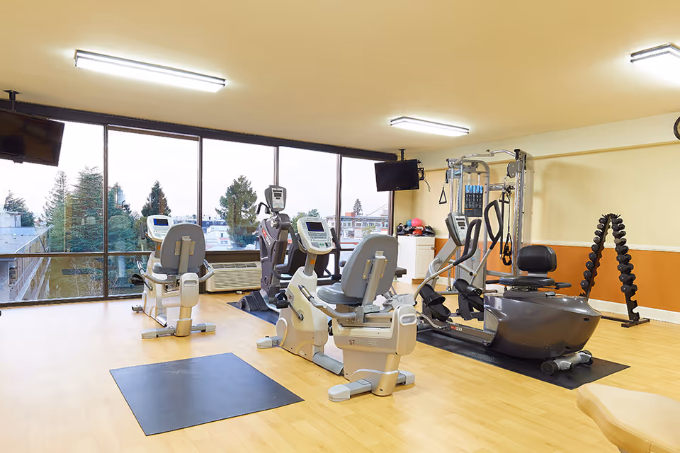 Bright senior living fitness room with recumbent bikes, an elliptical, strength machine and a dumbbell rack by large windows overlooking trees.