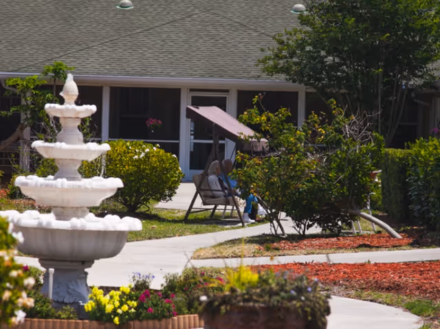 Courtyard with a three-tier stone fountain, flower beds, walkways, and two people seated on a shaded bench swing in front of a single-story building.