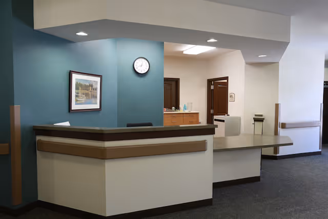 Reception desk and nurse station area inside an assisted living facility with a teal accent wall, wall clock, framed art, and adjacent hallway.