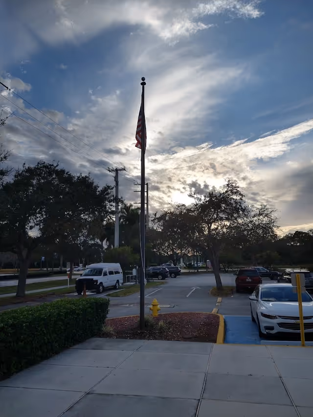 Flagpole with an American flag beside a small landscaped island and a parking lot under a cloudy sky.