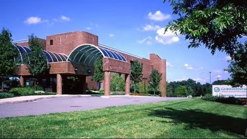 Exterior view of the Gurwin Jewish Nursing & Rehabilitation Center building with a brick facade, large glass canopy entrance, surrounded by green grass and trees under a blue sky with scattered clouds.