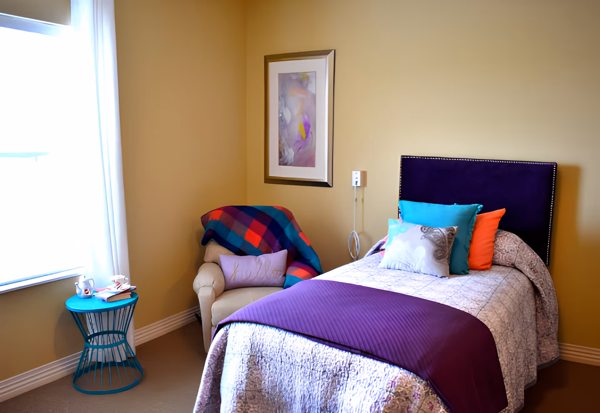 A cozy bedroom with a single bed featuring a purple headboard, a quilted bedspread, and several colorful pillows. Next to the bed is a beige armchair draped with a colorful plaid blanket and a lavender pillow. A small turquoise side table holds a cup, a book, and a pair of glasses. A framed abstract painting hangs on the wall above the armchair, and a window with white curtains lets in natural light.