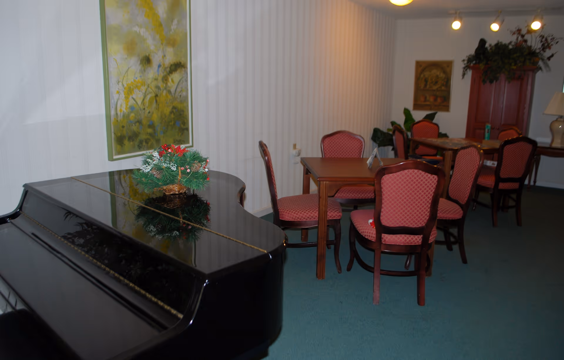 Interior room with a black grand piano on the left side and two wooden tables with red cushioned chairs arranged around them. There is a green carpet on the floor, a painting on the wall, and a red cabinet with plants on top in the background.