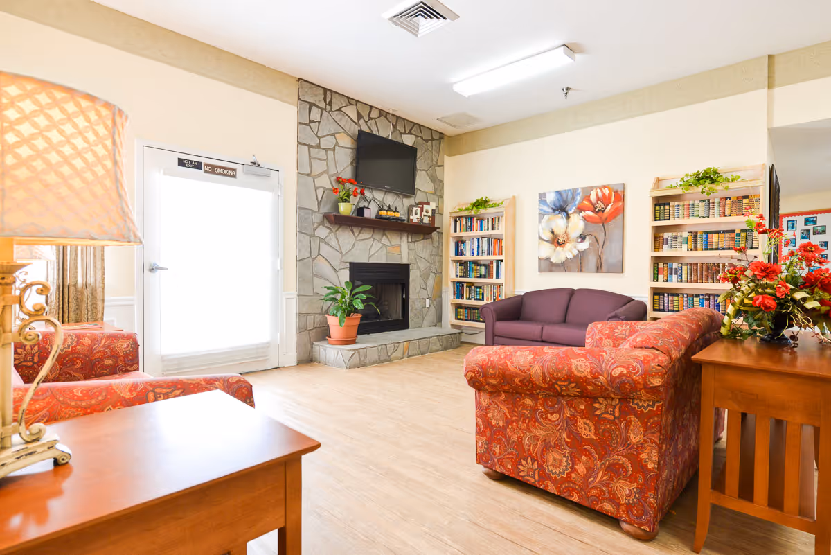 A cozy living room area with patterned red sofas, a purple loveseat, wooden bookshelves filled with books, a stone fireplace with a mounted TV above it, a potted plant, a wooden side table with a lamp, and a floral painting on the wall.