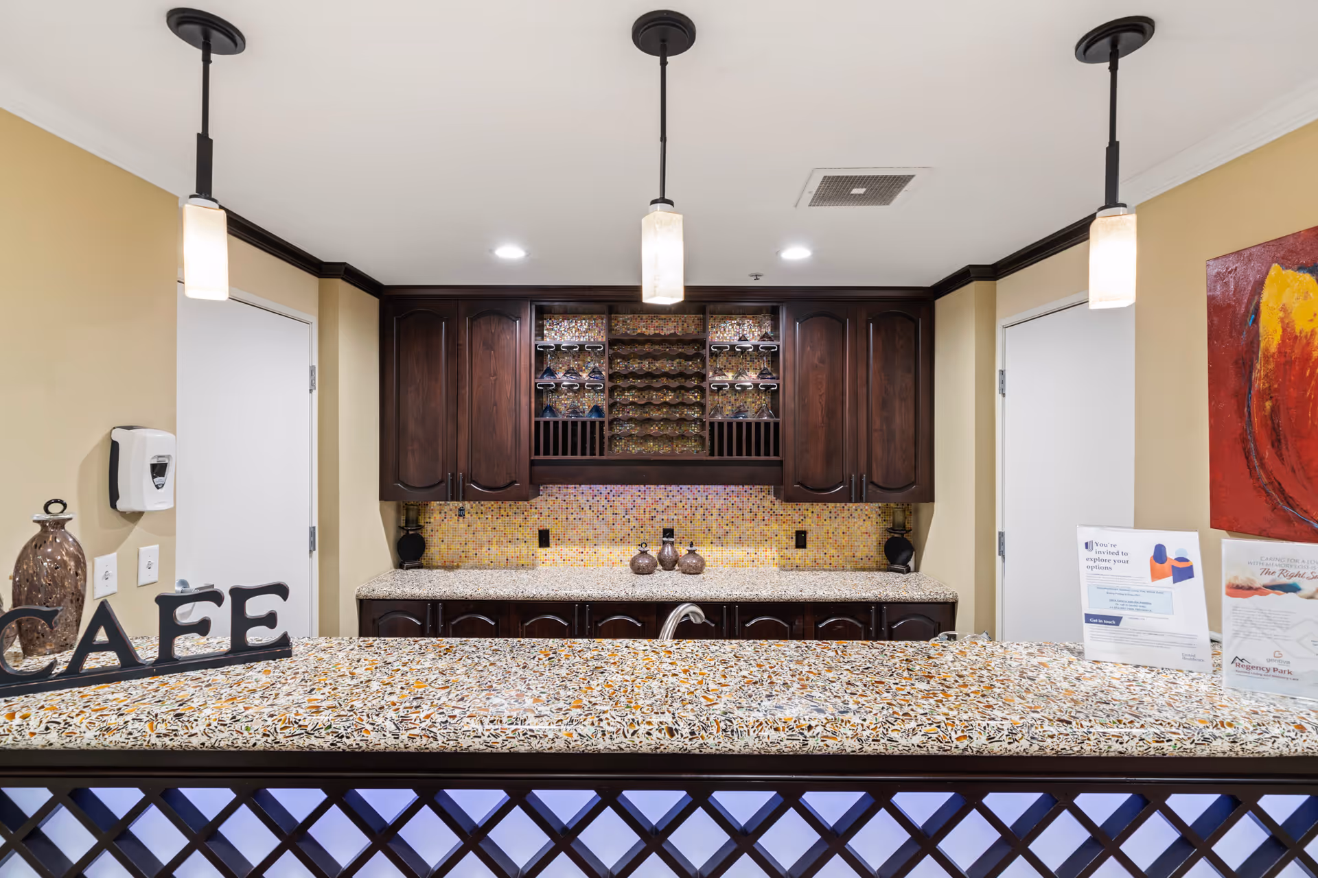 Interior view of a cafe counter area with a speckled countertop and dark wood cabinetry. The back wall features a wine rack with glasses and mosaic tile backsplash. Two pendant lights hang from the ceiling above the counter. There are decorative items on the counter including a sign that spells 'CAFE' and informational brochures. The walls are painted beige with white doors on either side of the cabinetry.