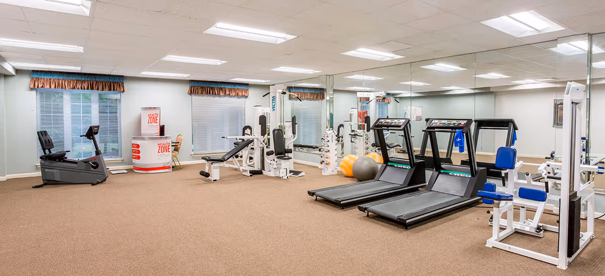 A fitness room with exercise equipment including two treadmills, a recumbent bike, weight machines, exercise balls, and dumbbells. The room has beige carpet, large mirrors on one wall, two windows with brown valances, and fluorescent ceiling lights.