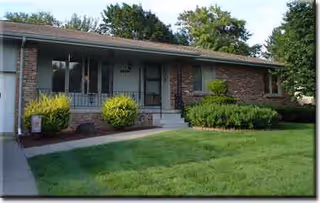 Single-story brick house with a manicured lawn, shrubs, and a front porch.