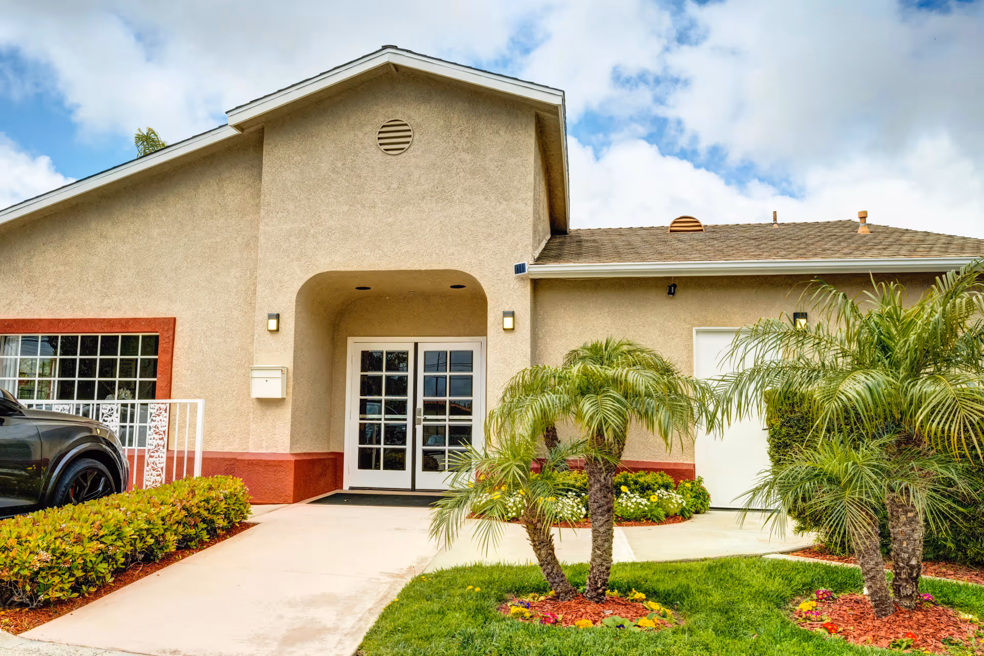 Exterior view of a beige stucco building with a red trim base, featuring a double glass door entrance under a covered archway. There are small palm trees and colorful flowers planted in mulched beds in front of the building, a black vehicle partially visible on the left, and a partly cloudy sky above.