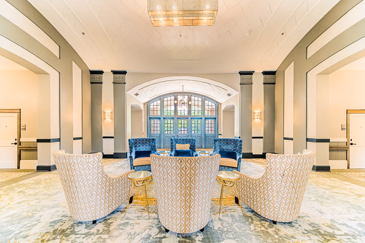 A bright and spacious senior living facility common area with a high ceiling and large arched windows. The room features a seating arrangement with three blue armchairs and three patterned beige armchairs surrounding small round tables. The walls are painted in neutral tones with decorative molding and sconces providing soft lighting.