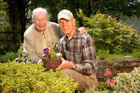 An elderly couple smiling and posing together in a garden. The woman is holding a basket with purple flowers, and the man is wearing a cap and plaid shirt. They are surrounded by green plants and flowers.