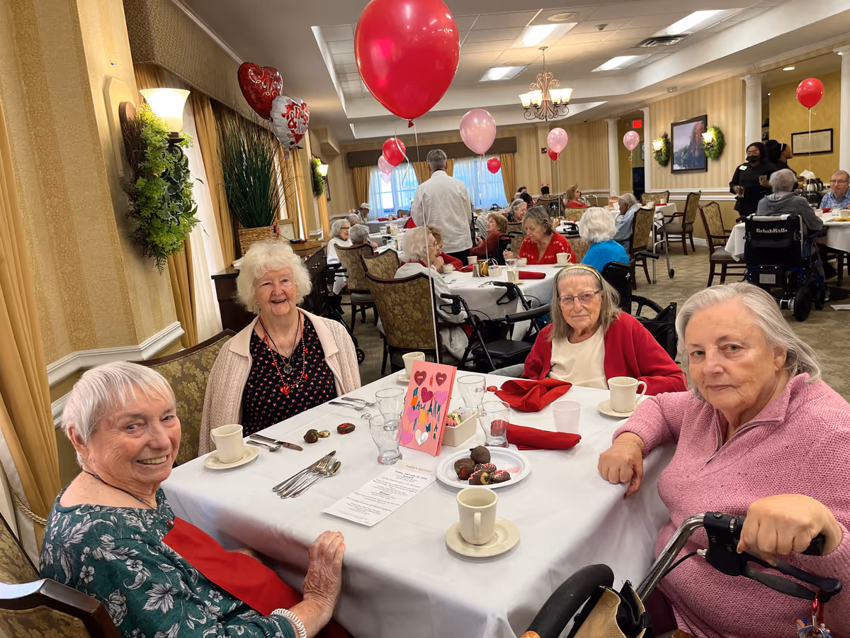 Four elderly women sitting around a dining table in a decorated room with red and pink balloons and heart-shaped decorations, enjoying a social gathering. The table is set with cups, plates with chocolates, and a Valentine's Day card. Other elderly people and staff are visible in the background.