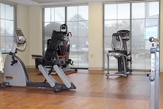 A fitness room with exercise equipment including a recumbent bike, an elliptical machine, and a treadmill, with large windows letting in natural light and a wooden floor.