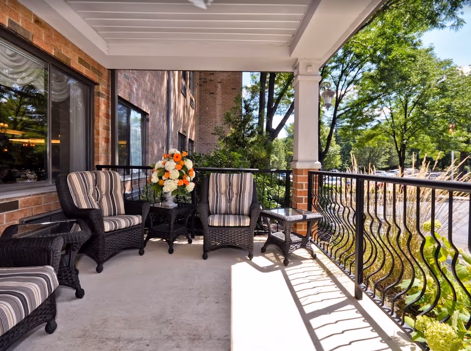 Outdoor covered patio area with black wicker chairs featuring striped cushions, a small glass-top table, and a flower arrangement with white and orange flowers. The patio has a black metal railing and overlooks a parking lot and green trees.