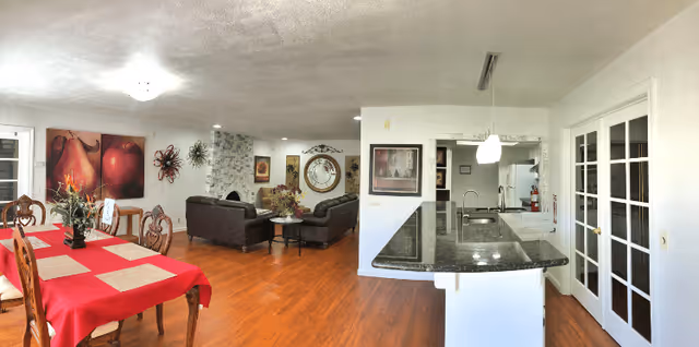 Interior view of a senior living facility showing a dining area with a table covered in a red tablecloth and wooden chairs, a living room with dark leather sofas and decorative wall art, and a kitchen area with a granite countertop and modern appliances. The space has wooden flooring and white walls with various framed pictures and a large decorative clock.