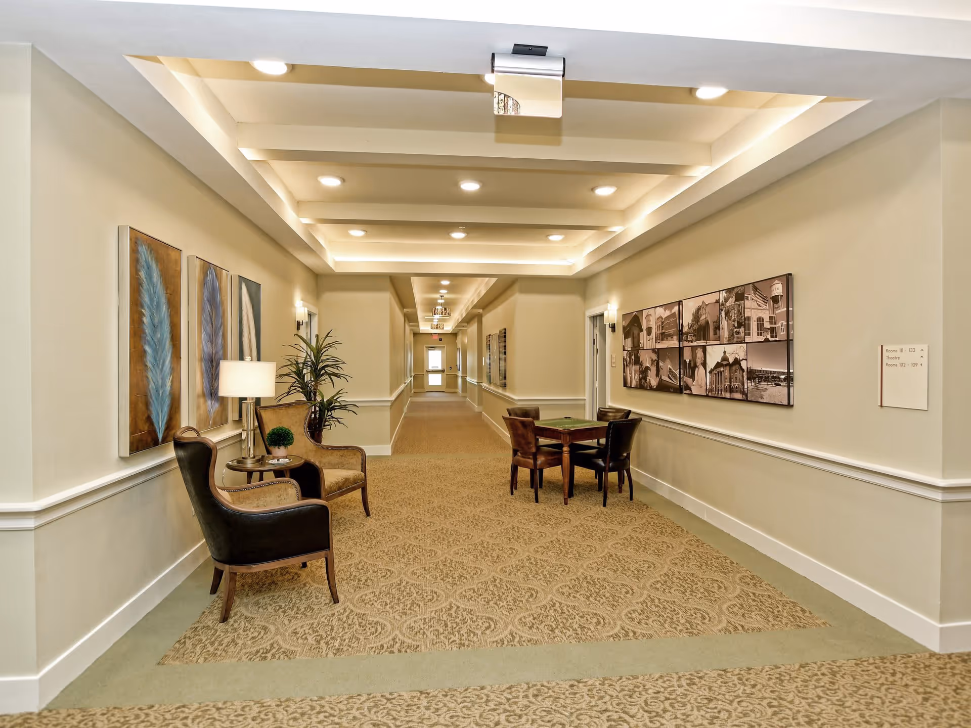 A well-lit hallway in a senior living facility with beige walls and patterned carpet. The hallway features a seating area with two armchairs, a small table with a lamp and plant, and a table with four chairs. Artwork and framed photos decorate the walls, and ceiling lights illuminate the space.