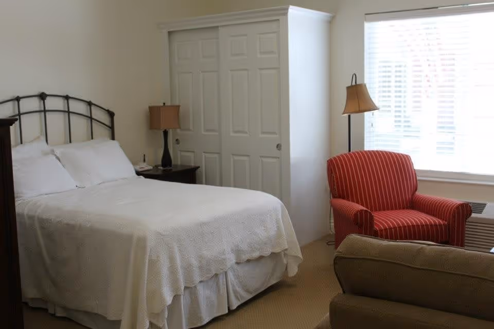 A bedroom with a neatly made bed covered in a white quilt, two white pillows, a black metal headboard, a dark wooden nightstand with a lamp, a red striped armchair near a window with white blinds, and a beige carpeted floor.