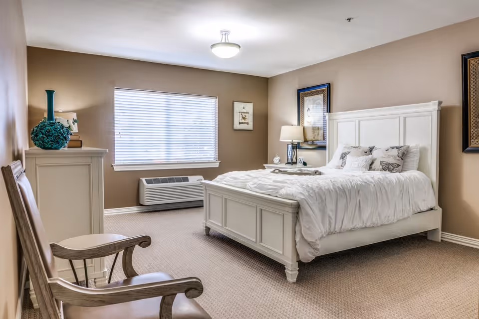 A cozy bedroom with beige walls and carpeted floor featuring a white wooden bed with white bedding and butterfly-patterned pillows. There is a wooden chair with armrests in the foreground, a white dresser with a decorative blue vase and books, a window with blinds, and framed artwork on the walls. A bedside table with a lamp and clock is next to the bed.