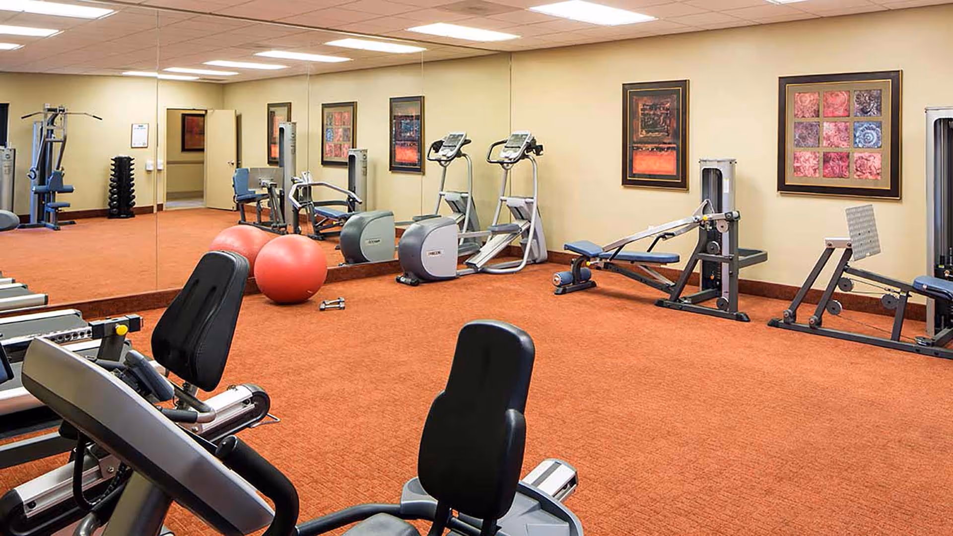 Interior view of a fitness room with exercise equipment including stationary bikes, elliptical machines, weight machines, and two large red exercise balls on a carpeted floor. The walls are decorated with framed artwork and a large mirror reflects part of the room.