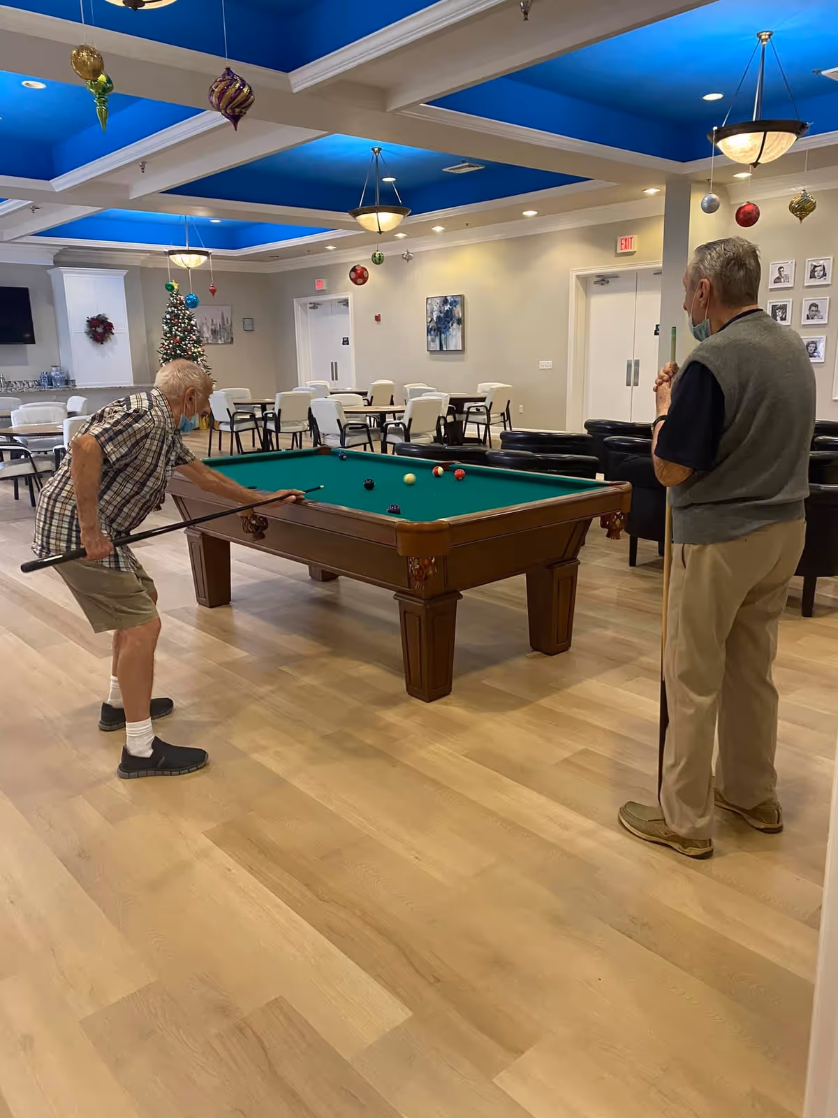 Two older men playing pool in a bright community recreation room with tables, chairs, and holiday decorations.