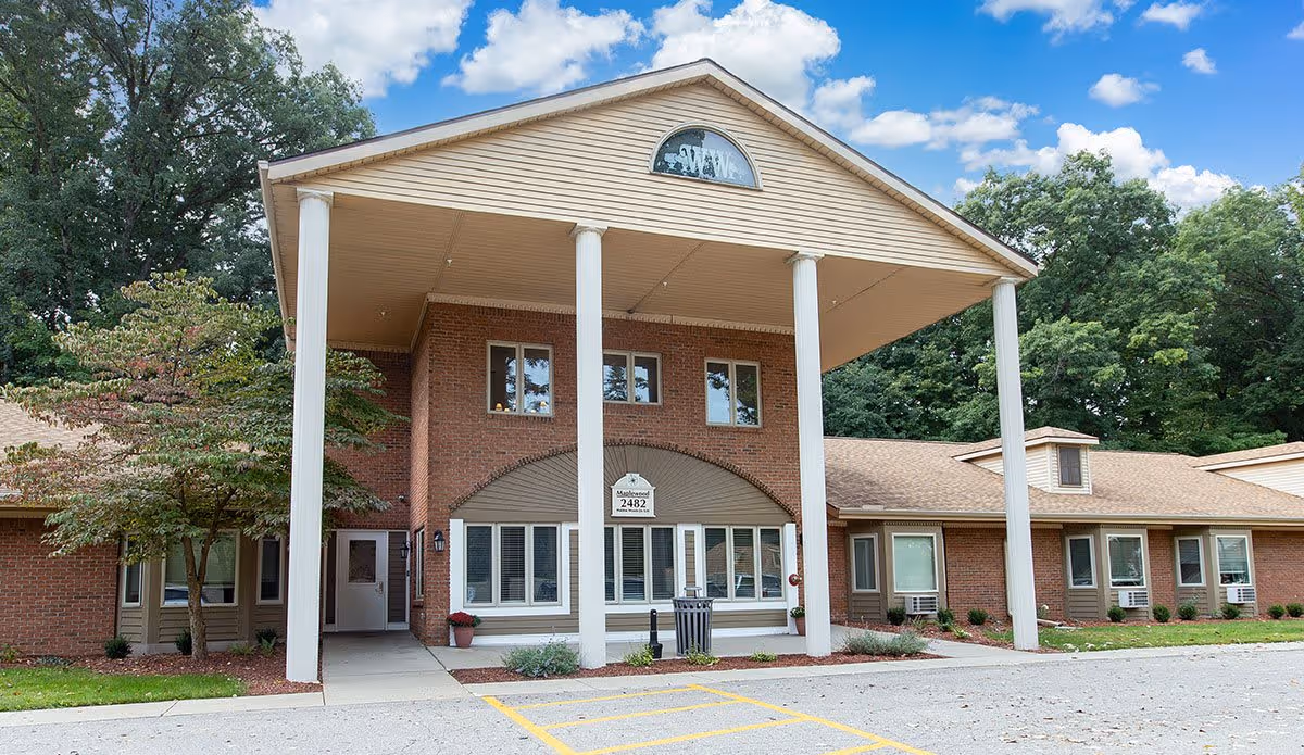 Front exterior view of a brick building with a large covered entrance supported by four white columns. The building has multiple windows, a sign above the entrance displaying the number 2482, and is surrounded by trees and a parking area with yellow lines.
