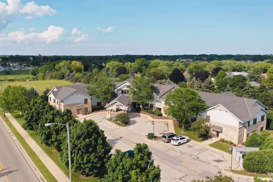 Aerial view of Riverview Village Senior Living facility showing multiple connected buildings with pitched roofs surrounded by trees and greenery. There is a driveway with parked cars and a sidewalk along the street. The sky is clear with some clouds.